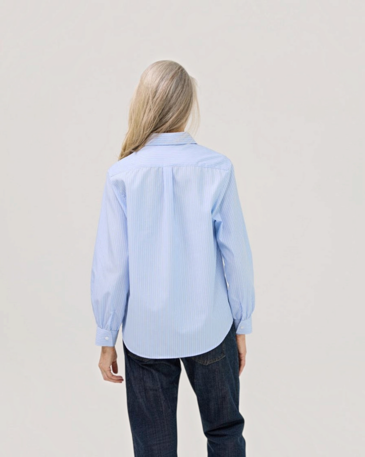 Back view of a woman wearing a Weldun blue and white striped cotton blouse with a curved hem and long sleeves.