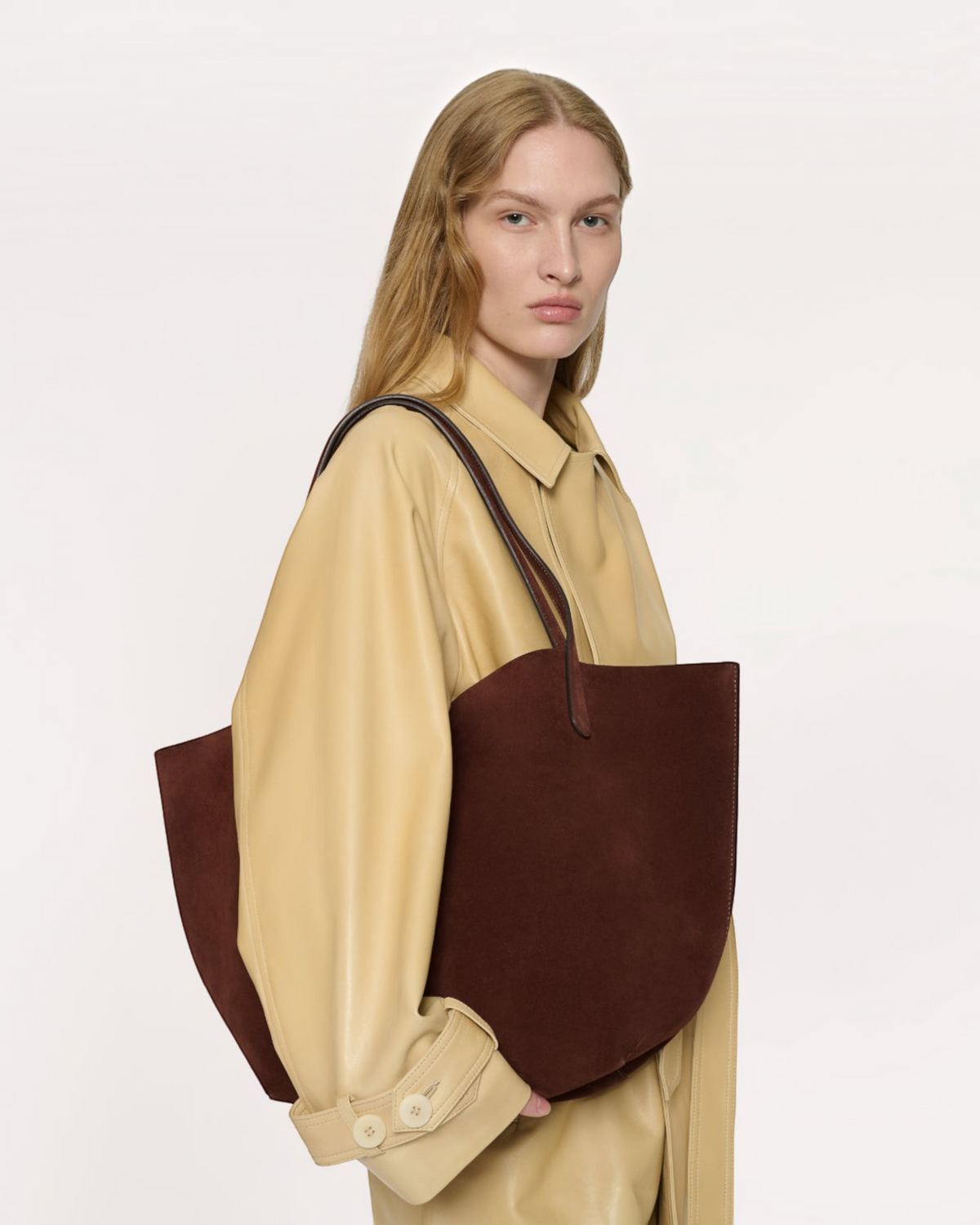 Weldun Mansur Gavriel chocolate brown suede tote bag carried on the shoulder by a model in a beige leather coat.