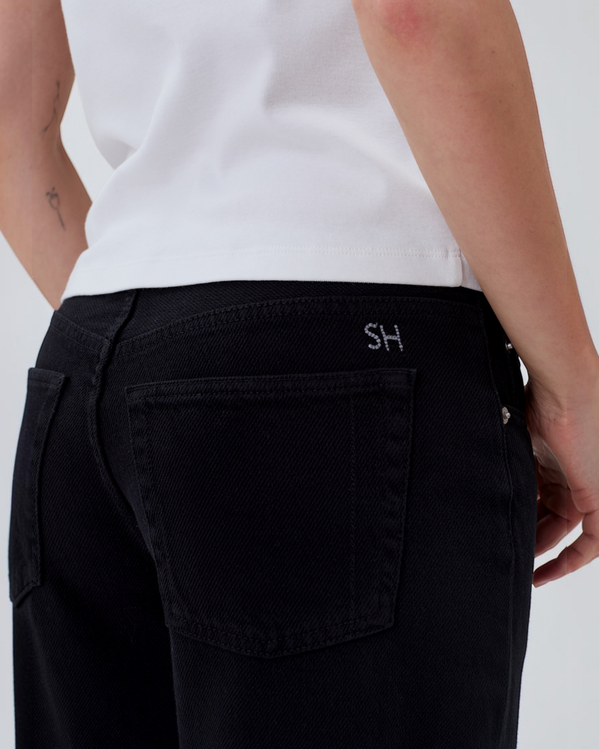 Weldun Still Here black relaxed fit jeans featuring back pockets and signature SH embroidery on a white t-shirt model