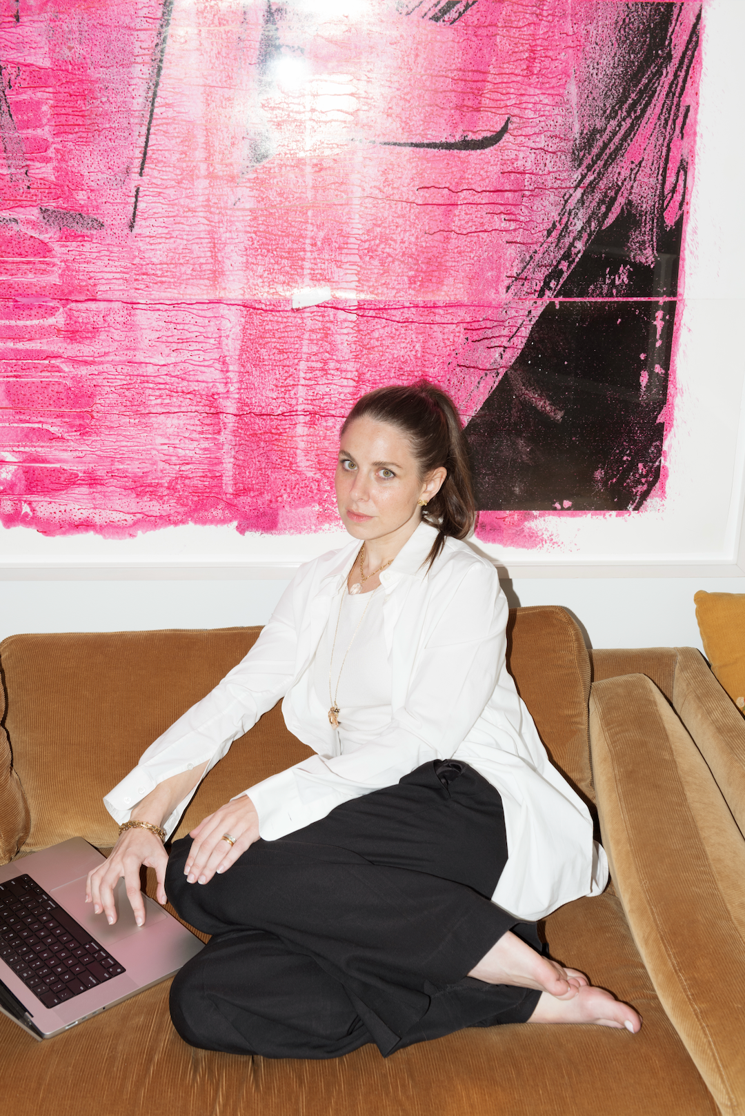 Ruthie Friedlander sits barefoot on a tan sofa with a laptop, in a white button-down and black trousers, in front of a large pink and black print.