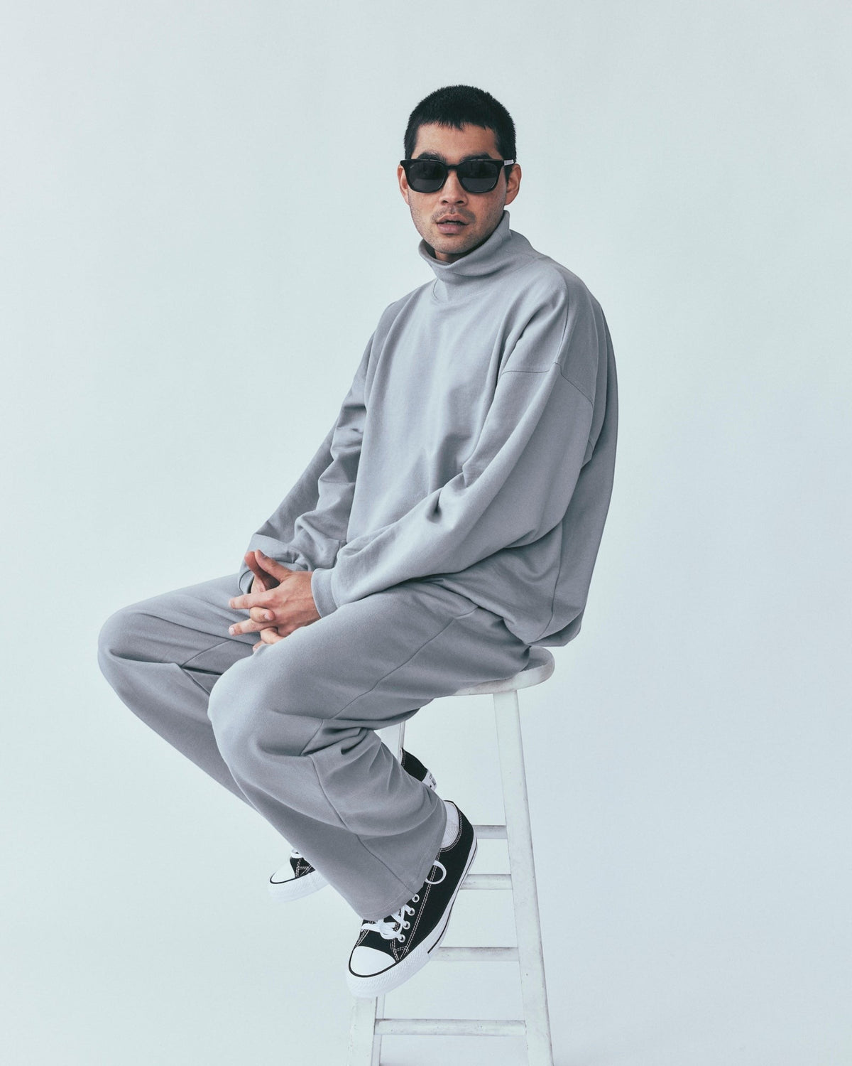 Weldun model wearing grey organic cotton sweatpants and matching turtleneck sweater while sitting on a white stool.