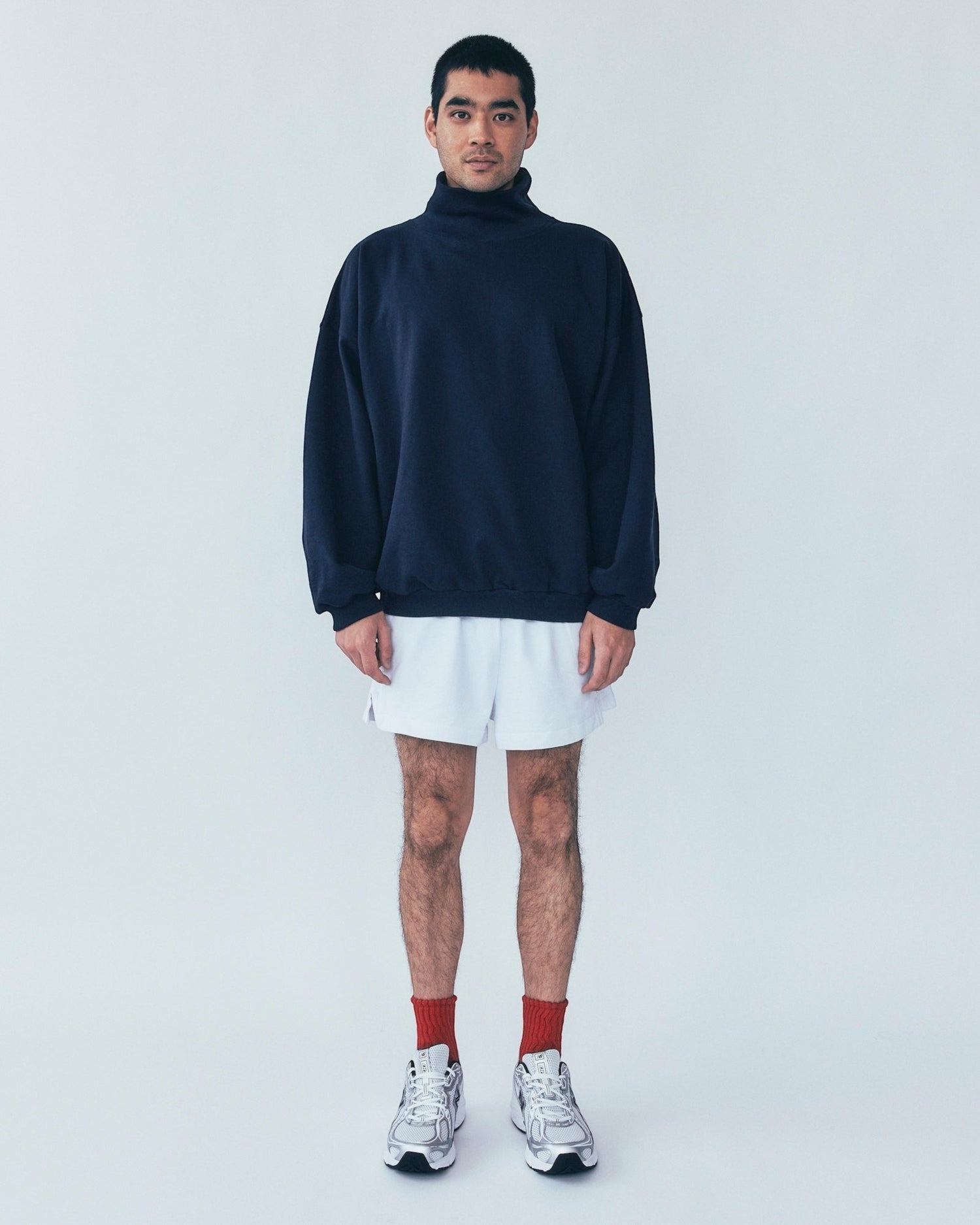 A model wearing a Weldun navy organic cotton high neck sweatshirt with a boxy fit, white shorts, and silver sneakers.