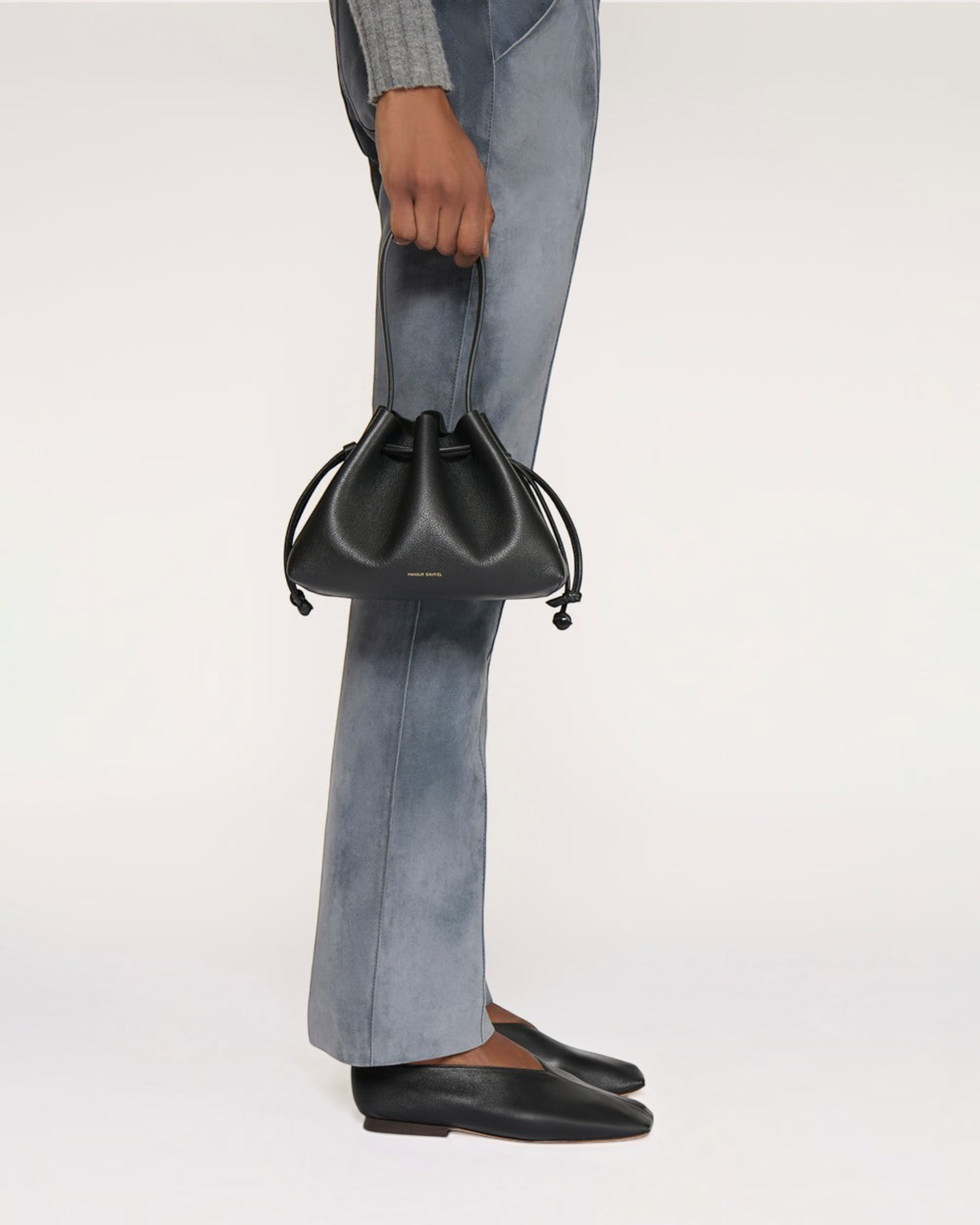 Weldun Mansur Gavriel black Italian leather drawstring pouchette held by a model wearing grey trousers.