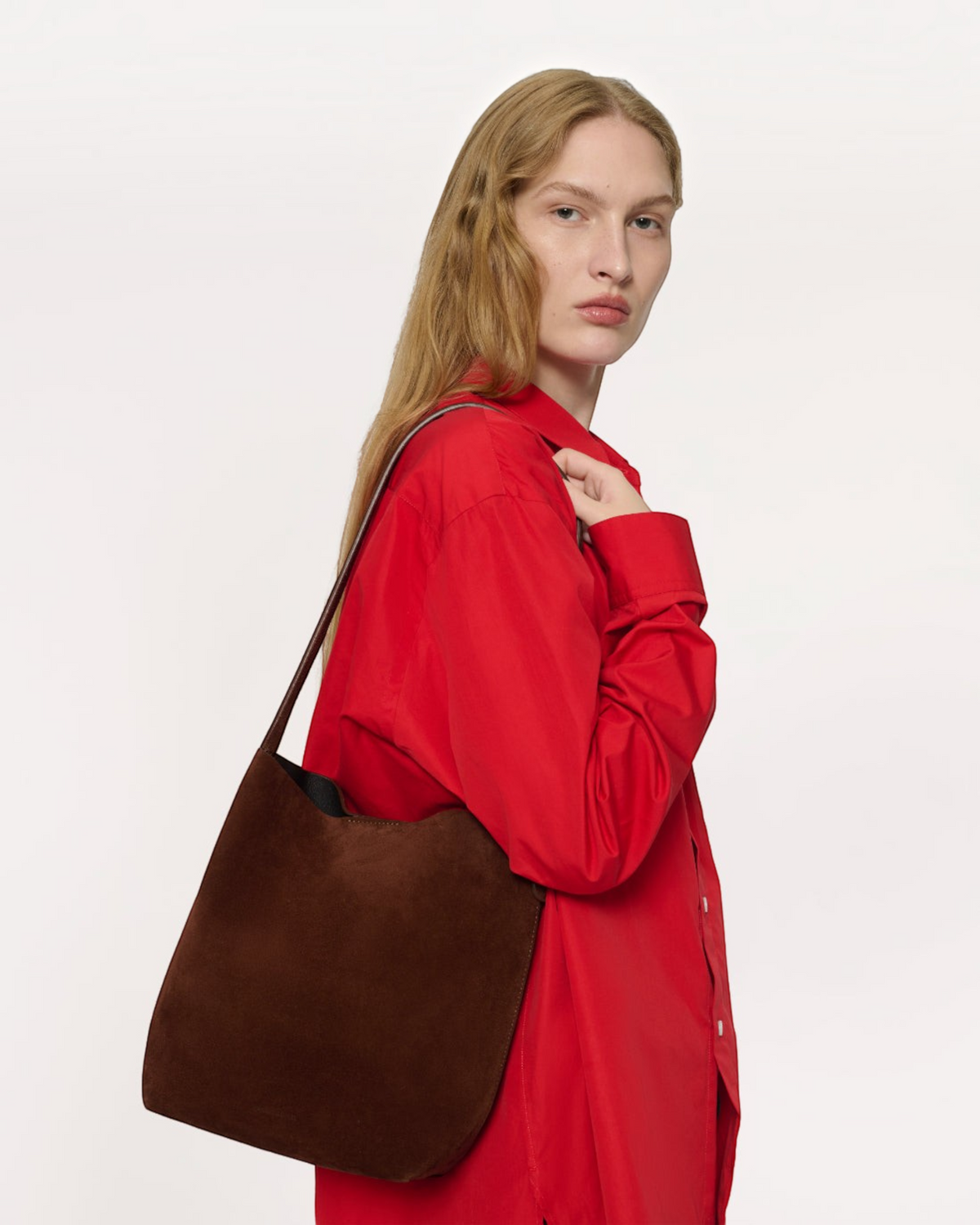 Weldun Mansur Gavriel Small Everyday Cabas in chocolate brown suede carried on the shoulder by a model in a red shirt.