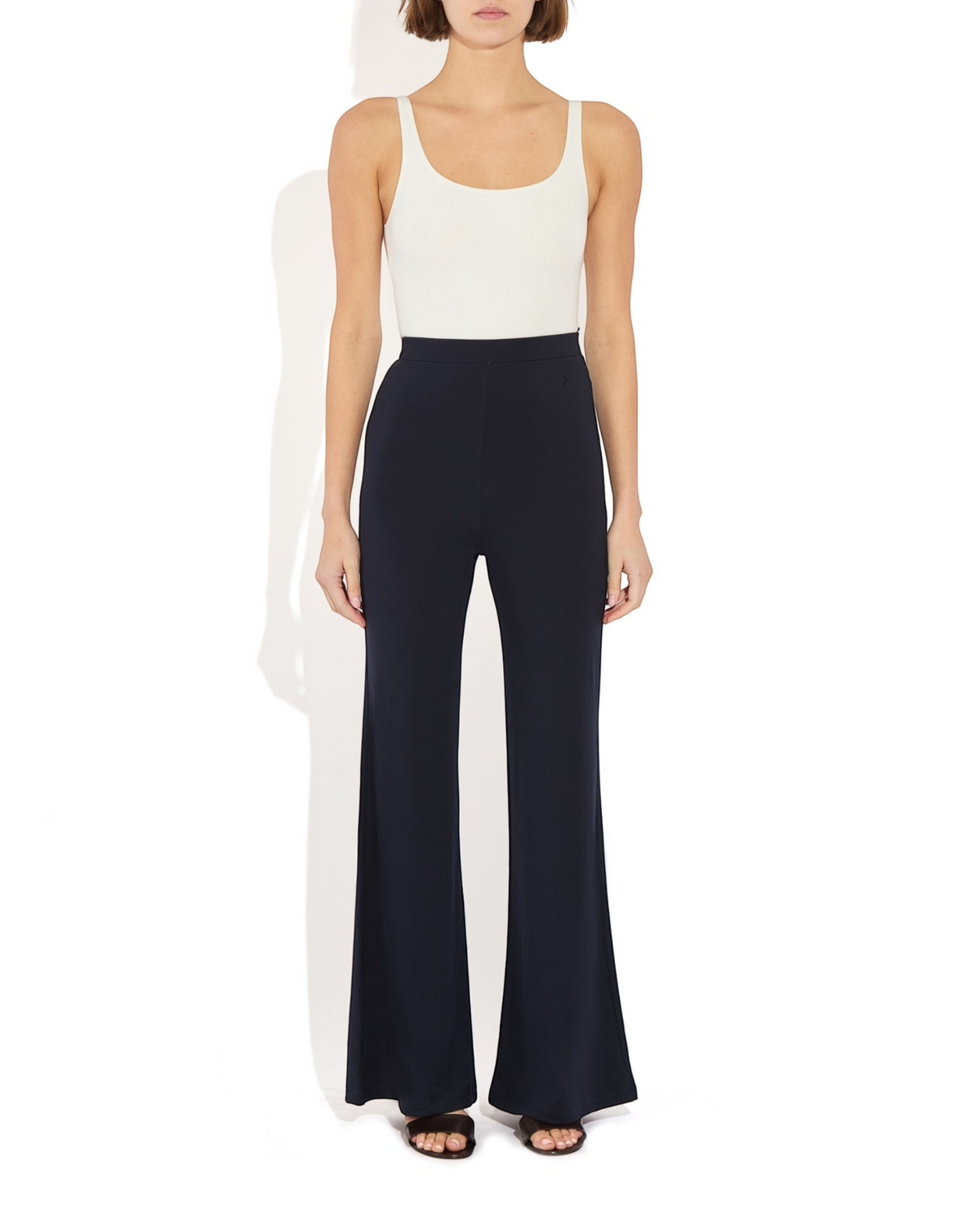 Model wearing a Weldun white scoop neck tank bodysuit tucked into high-waisted black flared trousers.