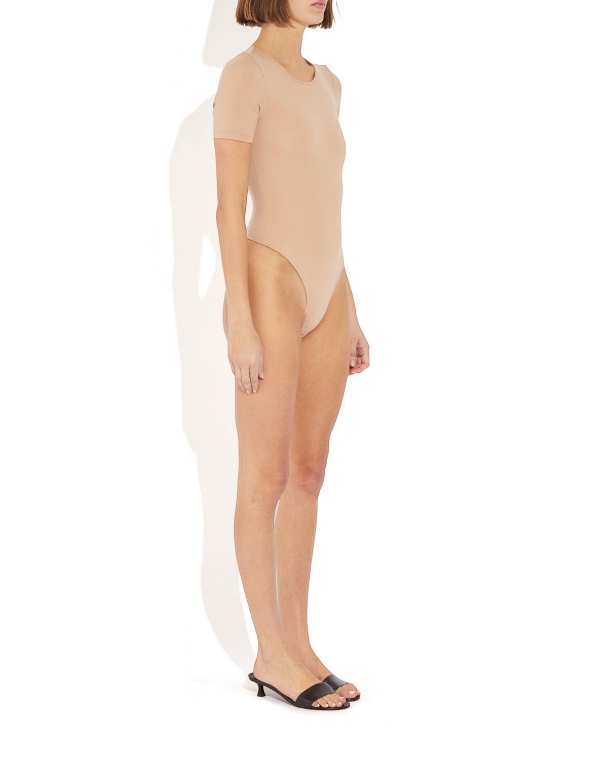 Model wearing a beige Weldun Penny short-sleeve bodysuit with a ballerina neckline and cheeky fit, side profile view.