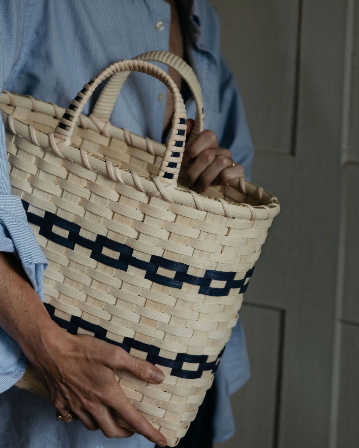 Weldun hand-woven rattan shopping basket with navy blue accents and wrapped handles held by a person in a blue shirt.