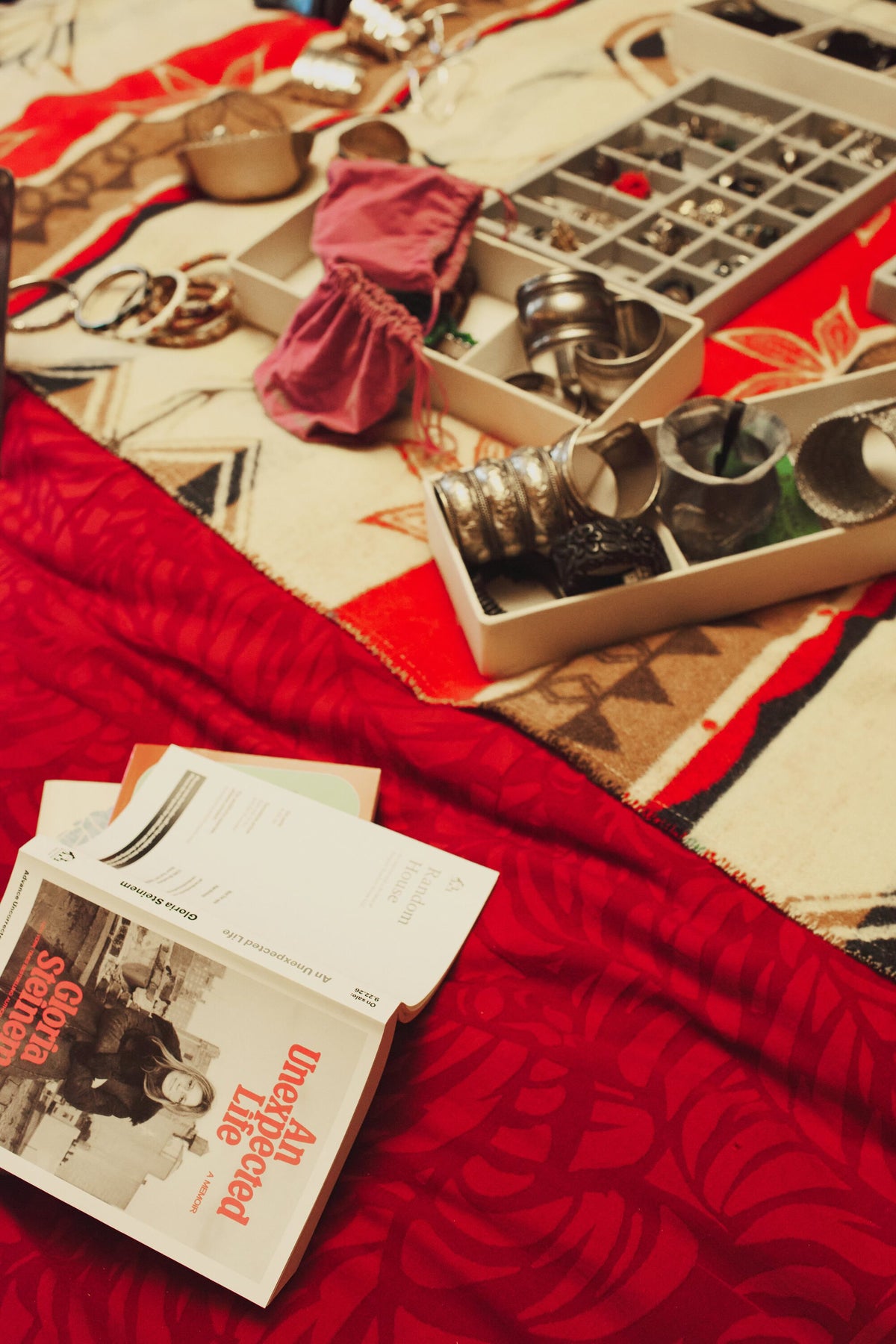 A tray of silver jewelry beside a copy of An Unexpected Life on a red blanket